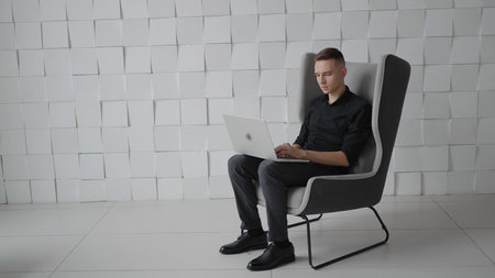 Young successful man in chair with laptop. Action. Young guy is working on laptop in business atmosphere. Business style chairs and rooms with successful manの写真素材