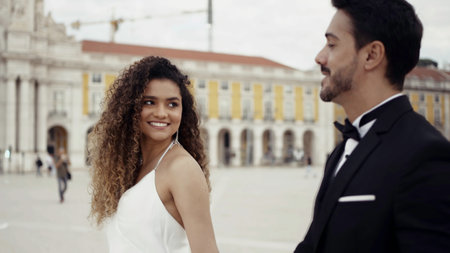 Young and beautiful couple walking along historic buildings and talking. Action. Man in suit and woman in white dress holding hands in the city square.の写真素材