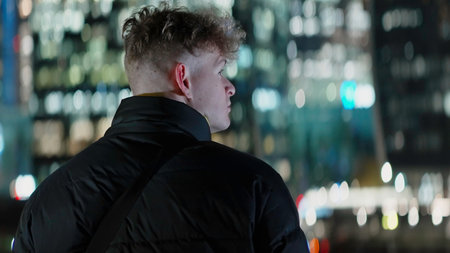 Man with short curly hair looking at the city at night. Action. Concept of travelling, man in warm jacket standing and looking at lights of building.の写真素材