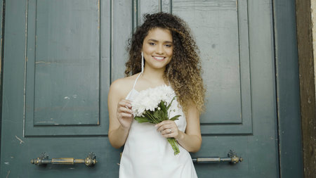 Gorgeous woman in white dress with curly hair with bouquet in her hands against the dark green door. Action. Bride waiting for the groom.の写真素材