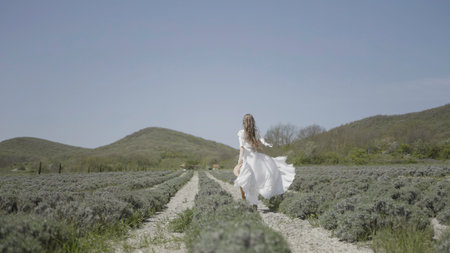 Rear view of a woman in white dress running in the field. Action. Running bride along farm fields on a summer day.の写真素材
