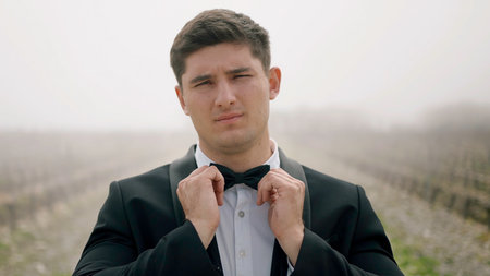 Young man in suit touching its bow tie outdoors. Action. Man wearing tuxedo standing in the field with fog.の写真素材