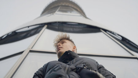 Bottom view of young man on background of tower. Action. Stylish young man in jacket at modern tower. Young man looks around warilyの写真素材