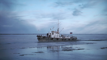 Fishing boat floats on background of cloudy winter landscape. Clip. Fishing boat with flag sails by ice in winter. Cloudy winter day with fishing boat on waterの写真素材