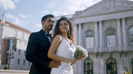 Elegant couple happily hugging. Action. Stylish couple in luxurious outfits hugging in city center. Newlyweds hug on background of ancient buildings on sunny summer dayの写真素材