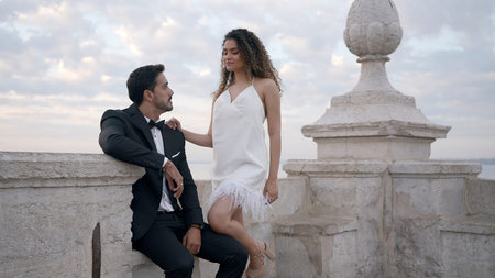 A young couple of models. Action. A woman in a short white dress walks along the pier with a man in a suit putting her long legs on him.の写真素材