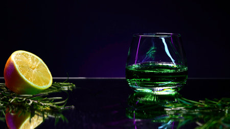 Close up of green alcoholic cocktail standing on a bar counter with orange and rosemary on a purple wall background. Stock clip. Concept of alcoholic addiction.の写真素材