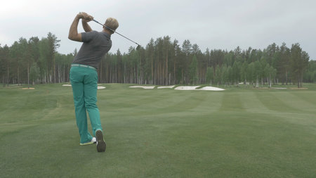Golfer Hitting Ball with Club on Beautiful Golf Course. Golfer hits an fairway shot towards the club house. Man hitting driver on a golf course in the sun. Professional male golf playerの写真素材