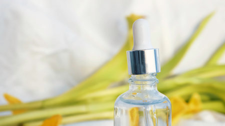 Vial with essential oil and yellow soft petals isolated on a white textile background. Girl hand touching cosmetic package surrounded by tulip leaves.の写真素材