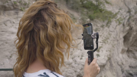 Beautiful woman takes pictures on phone with steadicam. Action. Young woman takes pictures on phone with manual stabilizer. Traveler takes pictures on hand-held steadicam in mountains.の写真素材