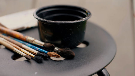 Close up of paint brushes with colorful stains and a small cup of water on a chair. Video. Concept...