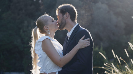Newlyweds kiss in nature with sunlight. Action. Beautiful couple of newlyweds in suits are kissing. Elegant couple of lovers kissing on background of sunlight and natureの写真素材