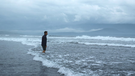 Man stands on water on seashore in cloudy weather. Clip. Lonely man stands in water with waves. Man...