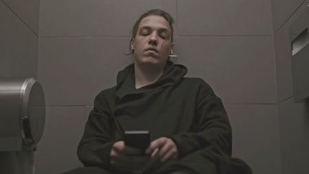 Young man with depression in toilet. Stock. Man sits on toilet and listens to sad music. Teenager...