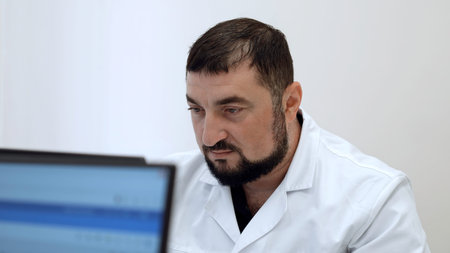 Middle aged doctor with in clinic. Clip. Bearded man in white coat looking at computer monitor.