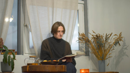 Young man is reading book and drinking tea in cafe. Media. Handsome young man is drinking tea and reading book alone. Guy is reading book in cozy cafeの写真素材