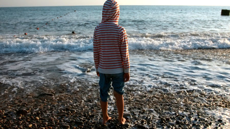Rear view of a boy in striped jacket standing on a pebble ocean shore. Creative. Child looking at waves on a sunny day.の写真素材