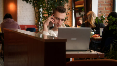 Young man is talking on phone on laptop. Stock footage. Handsome young man is talking on phone and working on laptop. Freelancer on remote with phone and laptop in cafeの写真素材