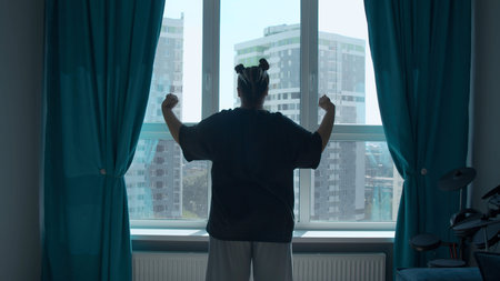 Rear view of a girl in casual clothes in modern bedroom in the morning. Media. Woman looking through...