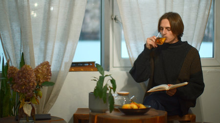 Young handsome man is reading book with tea in cafe. Media. Student is sitting alone and reading book in cozy cafe. Young man is reading book and drinking tea in student cafeの写真素材