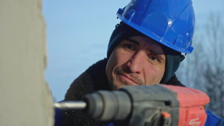 Worker in hard hat drills wall with drill. Clip. Man drills concrete walls at construction site. Man drills walls with drill in house under constructionの写真素材