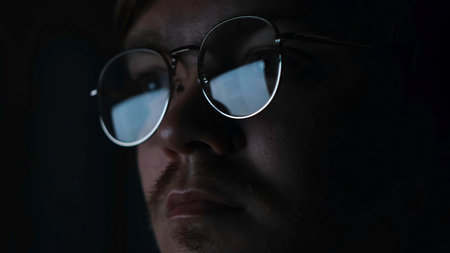 Close up of serious focused man scientist face in glasses looking at screen in the dark room. Concept. Male reading information on computer monitor that is reflected in glasses.の写真素材