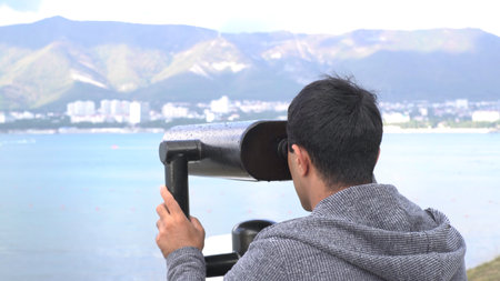 A man looking through coin binoculars on fantastic mountain and sea view. Media. Male tourist looks at the coastal city through coin binoculars.の写真素材