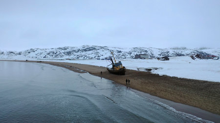 Aerial view of the old rusty ships left on the sandy shore by the ocean shore in winter season. Footage. Flying over people walking on the beach near cold ocean and snowy hills.の写真素材