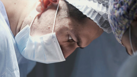 Close up of male surgeon face at work. Action. Side view of a concentrated doctor looking down and wearing protective medical uniform, concept of health.の写真素材