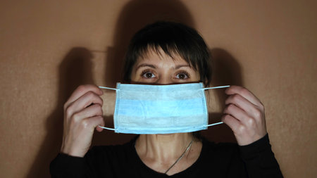 Front view of woman putting onprotective mask. Concept. Portrait of dark haired female wearing blue medical mask, standing in front of the beige wall, covid 19.の写真素材
