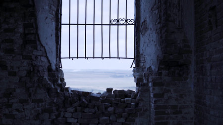 Window frame looking towards small village, green fields and blue cloudy sky. Footage. View from the ruined building with a destroyed window with iron lattice.の写真素材