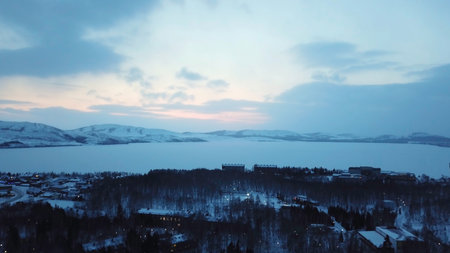 Aerial dramatic winter landscape over city covered by white snow in winter season in the evening. Flying over frozen white lake, trees and buildings on blue cloudy sky background.の写真素材