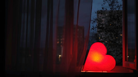 Nightlight in the shape of a heart in the children room. Concept. Red beautiful heart shaped night lamp turned on while standing on windowsill in front of window.の写真素材