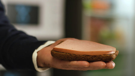 Delicious sausage sandwich. Stock footage. Close-up of mans hand holding a piece of Borodino rye bread with slice of sausage falling on it.の写真素材