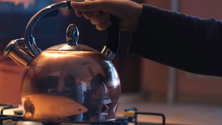 Woman placing teapot on the gas stove in the kitchen. Concept. Close up of young female reflected in the polished surface of steel kettle putting it on a turned off gas stove.の写真素材