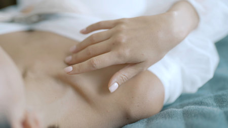 Close up of a young woman touching gently her collarbones while lying on bed in white dress. Action. Tender female relaxing on bed.の写真素材