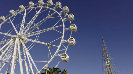 White ferris wheel rotating under blue sky near a sailing ship. Actionの写真素材