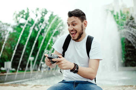 Cheerful handsome man sitting and reviewing photos from camera on fountains background.