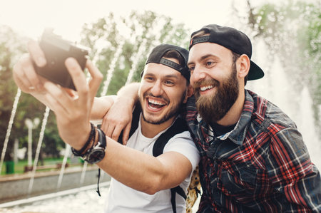 Image of two hipsters with beard in public park smiling and taking a selife with digital camera.の写真素材