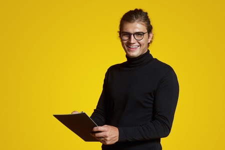 Attractive young joyful businessman with eyeglasses, wearing turtleneck, looking at camera and holding a notepad isolated on yellow backgroundの写真素材