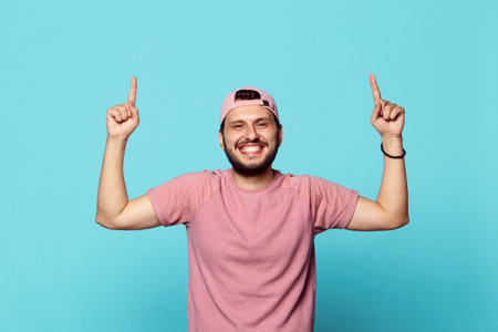 Young handsome man wearing pink tshirt and baseball cap, pointing fingers up at copyspace isolated...