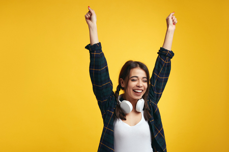 Laughing young girl in shirt and white headphones on neck, holding hands up in gesture of victory, closes eyes from pleasure, on yellow background.の写真素材