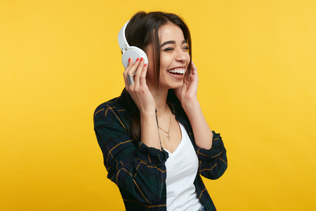 Overjoyed young girl enjoy sound of music, keeps hands on headphones, closes eyes from pleasure, listens song and feels glad, expresses happiness. Cute female wearing stylish outfit above yellow wallの写真素材
