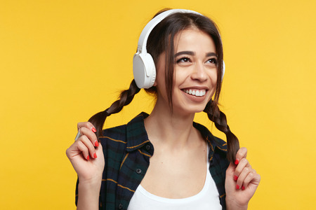 Cheerful young girl in stylish clothes and white headphones, listens music and feels glad, playing with her pony tails, looking away with smile, isolated over yellow backgroundの写真素材