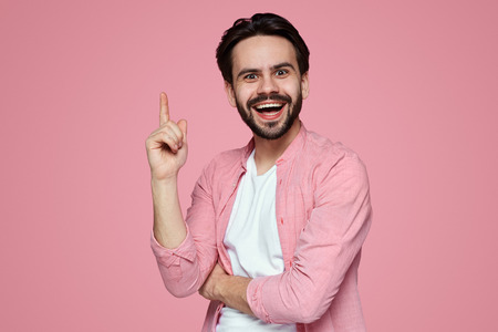 Studio shot of intelligent bearded man raises fore finger as recieves good idea or glad find out about successfully passed exam. Excited cheerful man remembers something pleasant, isolated on pinkの写真素材