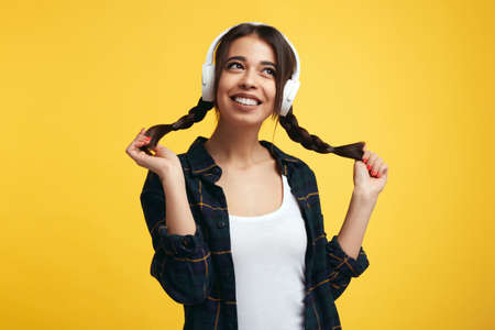 Cheerful young girl in stylish clothes and white headphones, listens music and feels glad, playing with her pony tails, looking away with smile, isolated over yellow backgroundの写真素材