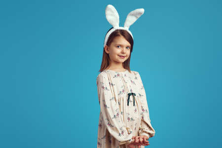 Pretty little girl in white shirt and bunny ears posing adorably on blue background smiling at cameraの写真素材