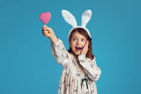 Beautiful girl kid smiling and holding tasty sweet heart shape cookie, wearing bunny ears, isolated over blue wallの写真素材