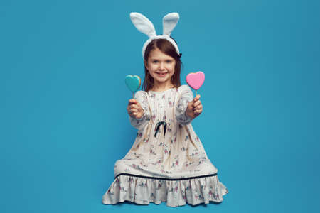 Nice cute kid girl having two cookies in heart shape on sticks, wearing easter bunny ears, isolated over blue backgroundの写真素材
