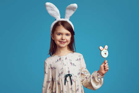 Adorable charming girl smiling and holding a cookie in shape of rabbit, wearing bunny ears over blue wallの写真素材